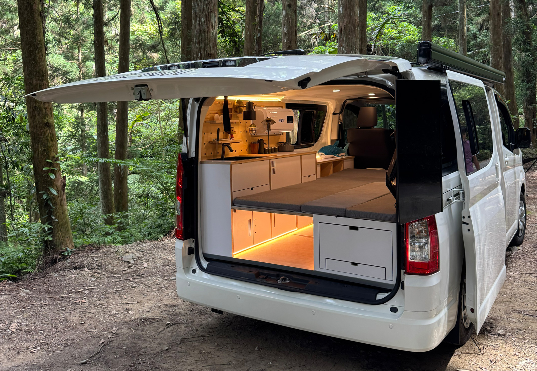 野孩子 Hiace 海力士露營車停在森林中，車尾展開床鋪、白色木作櫃體與暖色燈光