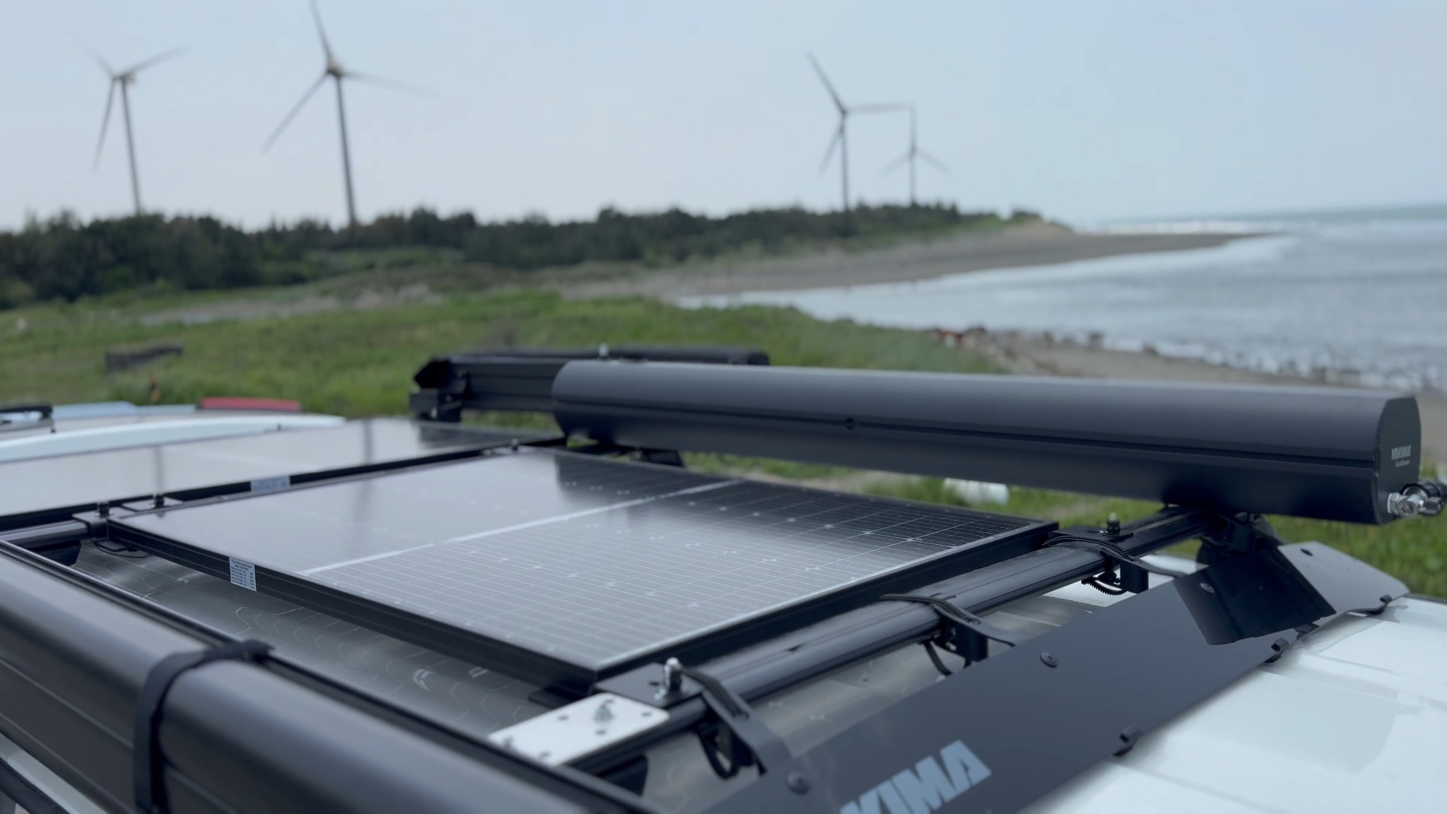 露營車車頂太陽能板與架體近照，背景為海岸與風力發電機
